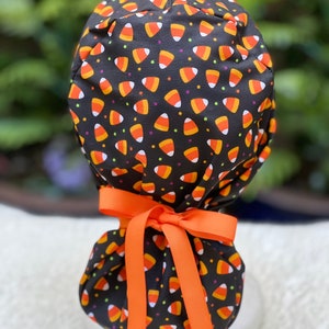 May include: A black scrub cap with a candy corn pattern in orange, yellow, and white. The cap is tied with an orange ribbon bow. The background is blurred green foliage.