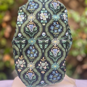 May include: A patterned scrub cap featuring a dark navy background with a repeating floral design in shades of green, blue, and cream. The cap has a rounded shape and is made of fabric. The floral pattern includes stylized flowers and leaves.