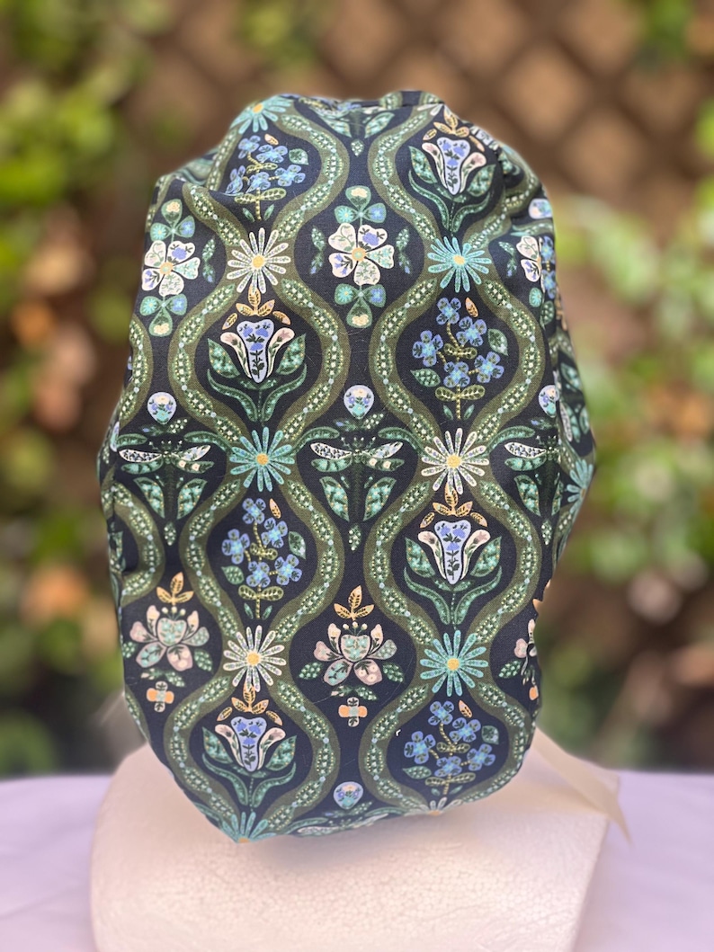 May include: A patterned scrub cap featuring a dark navy background with a repeating floral design in shades of green, blue, and cream. The cap has a rounded shape and is made of fabric. The floral pattern includes stylized flowers and leaves.