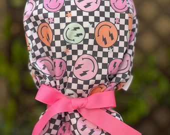 Pode incluir: Uma touca estampada com fundo xadrez preto e branco, com rostos sorridentes e raios em rosa, laranja, verde e azul claro. A touca é amarrada com um laço rosa brilhante.