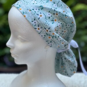 K&ouml;nnte beinhalten: Eine hellblaue OP-M&uuml;tze mit wei&szlig;em Blumenmuster. Die M&uuml;tze hat einen Bindegurt am Hinterkopf.