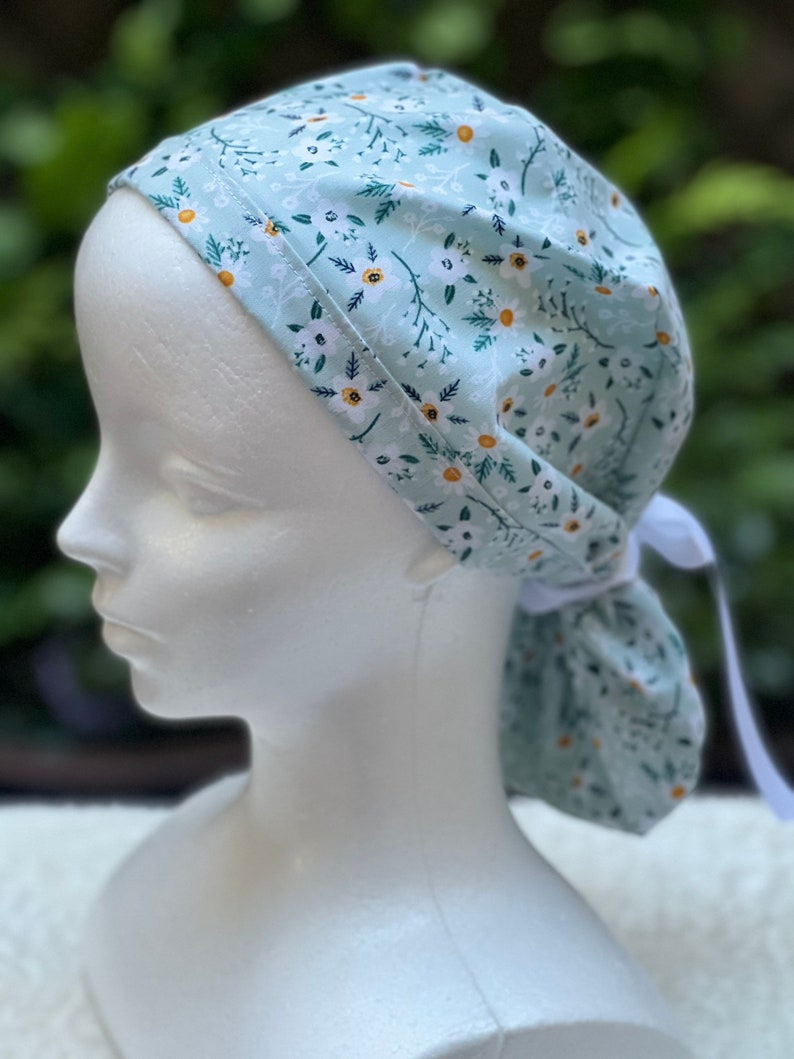 K&ouml;nnte beinhalten: Eine hellblaue OP-M&uuml;tze mit wei&szlig;em Blumenmuster. Die M&uuml;tze hat einen Bindegurt am Hinterkopf.