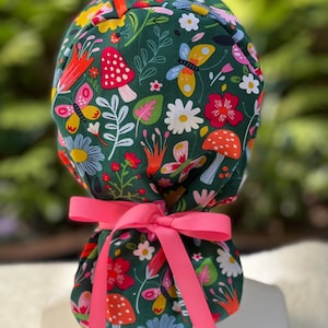 May include: A dark green scrub cap with a colorful floral pattern featuring mushrooms, butterflies, and flowers. The cap is tied with a bright pink ribbon. The design includes red, yellow, and white accents.