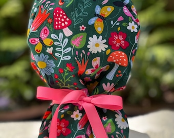 May include: A dark green scrub cap with a colorful floral pattern featuring mushrooms, butterflies, and flowers. The cap is tied with a bright pink ribbon. The design includes red, yellow, and white accents.