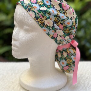 May include: A surgical scrub hat with a floral print featuring pink, white, and yellow daisies on a dark green background. The hat has a pink tie.