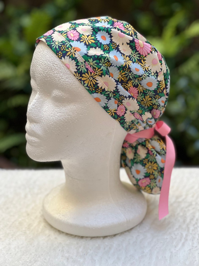 May include: A surgical scrub hat with a floral print featuring pink, white, and yellow daisies on a dark green background. The hat has a pink tie.