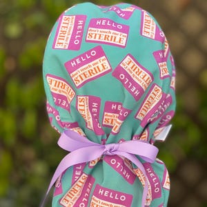 Peut inclure: Un bonnet de chirurgien turquoise avec un motif répété d'étiquettes roses indiquant "HELLO don't touch me I'm STERILE". Le bonnet est noué avec un ruban lavande. Cet accessoire médical est destiné aux professionnels de la santé.