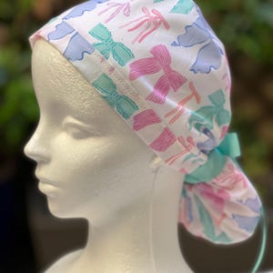 May include: A white surgical scrub hat with a colourful bow pattern. The hat has a turquoise tie and is made of soft, breathable fabric.