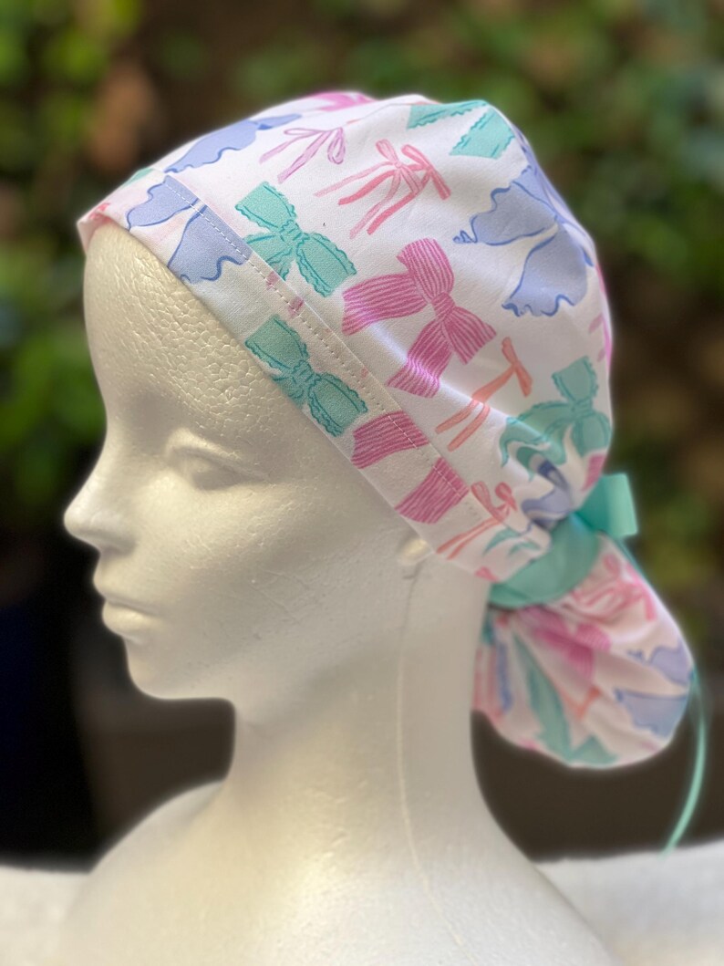 May include: A white surgical scrub hat with a colourful bow pattern. The hat has a turquoise tie and is made of soft, breathable fabric.