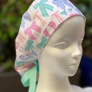 May include: A white surgical scrub hat with a colourful bow print. The hat has a green tie that can be tied at the back of the neck.