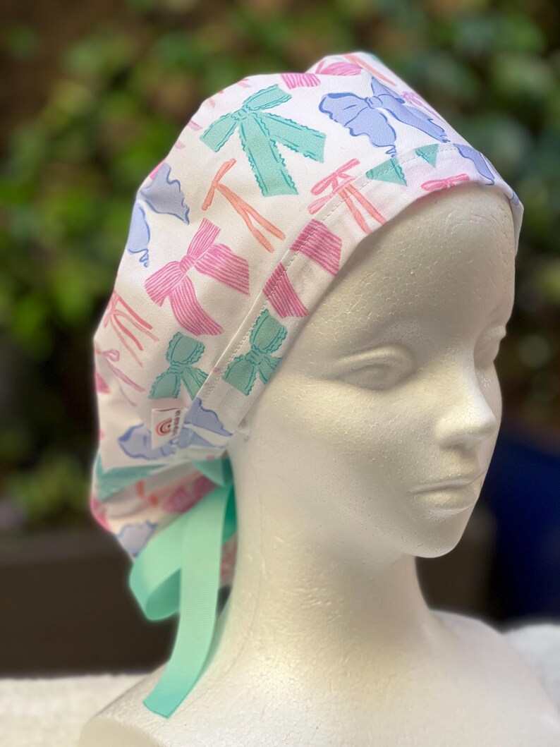 May include: A white surgical scrub hat with a colourful bow print. The hat has a green tie that can be tied at the back of the neck.
