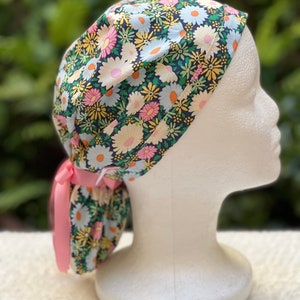 May include: A black scrub hat with a floral print featuring white, yellow, and pink daisies. The hat has a pink ribbon tie.
