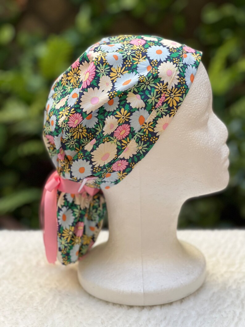 May include: A black scrub hat with a floral print featuring white, yellow, and pink daisies. The hat has a pink ribbon tie.