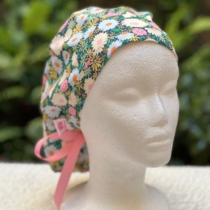 May include: A floral print surgical scrub hat with a pink tie. The hat has a dark blue background with white, pink, and yellow flowers.