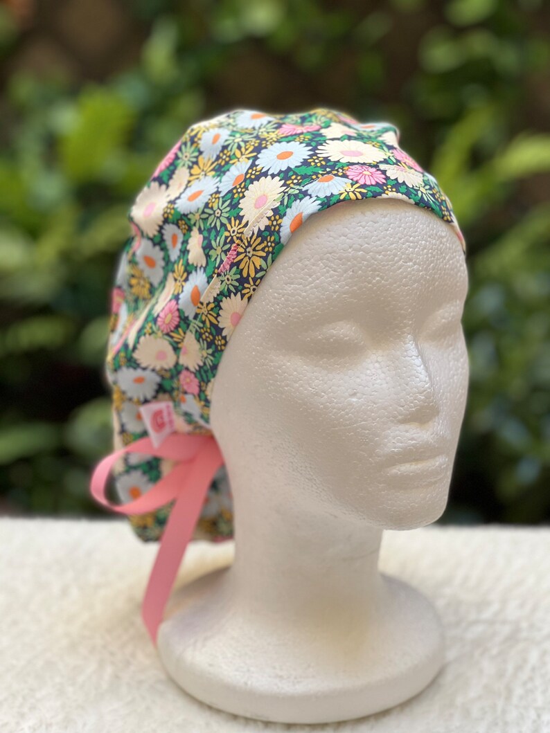 May include: A floral print surgical scrub hat with a pink tie. The hat has a dark blue background with white, pink, and yellow flowers.
