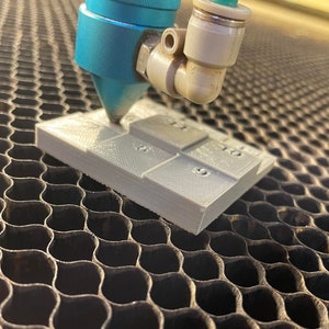 Puede incluir: Primer plano de una máquina de corte láser en funcionamiento. El cabezal láser azul se enfoca en un pequeño bloque rectangular gris claro con cuadrados numerados. El bloque está sobre una superficie metálica de panal.