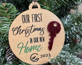May include: A round wooden Christmas ornament with the text "Our First Christmas in Our New Home 2023". A red glitter key is attached to the ornament.