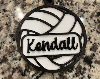 May include: A white and black wooden volleyball ornament with the name "Kendall" written on it. The ornament is hanging from a black cord with white and pink beads.
