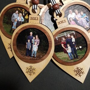 May include: Five wooden Christmas ornaments with a snowflake design. Each ornament has a photo frame with a family photo and the year 2022 engraved on the top. The ornaments are attached to black and white striped ribbon.