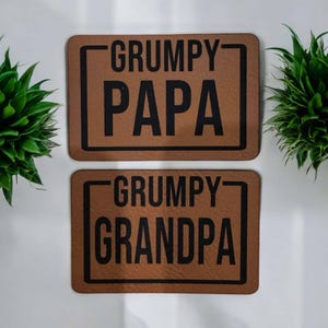 Puede incluir: Dos parches rectangulares de cuero marrón con texto negro. El parche superior dice "GRUMPY PAPA" y el parche inferior dice "GRUMPY GRANDPA". Cada parche tiene un borde negro sobre un fondo blanco.