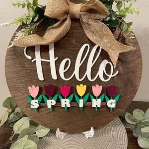 May include: A round wooden sign with the words "Hello Spring" in white letters. The sign is decorated with a burlap bow and green leaves. There are also small wooden tulips in various colors around the bottom of the sign.