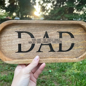 Può includere: Vassoio ovale in legno chiaro. La parola "DAD" è incisa in nero, con il messaggio "YOU ARE THE PIECE THAT HOLDS THIS FAMILY TOGETHER" sotto. Un regalo perfetto per la festa del papà.