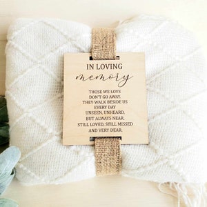May include: A white knitted throw blanket with a wooden sign attached. The sign has the text "IN LOVING memory THOSE WE LOVE DON'T GO AWAY. THEY WALK BESIDE US EVERY DAY UNSEEN, UNHEARD, BUT ALWAYS NEAR, STILL LOVED, STILL MISSED AND VERY DEAR."