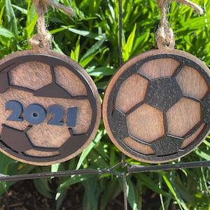 May include: Two wooden ornaments shaped like soccer balls with black outlines. One ornament has the year 2021 printed on it in blue.