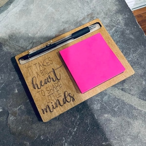 Puede incluir: Organizador de escritorio de madera con un bolígrafo, un bloc de notas rosa brillante y la frase grabada "It takes a big heart to shape little minds". El organizador es de madera clara.