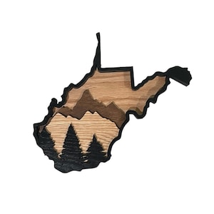 May include: A layered wooden wall art piece in the shape of West Virginia. The artwork features a black outline, with a natural wood grain interior. The design includes mountain and tree silhouettes, creating a three-dimensional effect. The background is white.