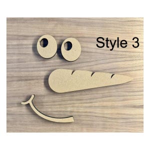 May include: Wooden snowman face cutouts, including two eye circles, a carrot nose with etched lines, and a curved smile. The text "Style 3" is printed in black. The cutouts are made of light brown wood and are arranged on a wooden surface.