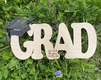 Könnte beinhalten: Ein Holzschild mit dem Wort "GRAD" in großen, stilisierten Buchstaben. Eine schwarze Studentenmütze ist an der oberen linken Ecke des Schildes befestigt. Das Schild enthält auch den Text "Denn ich weiß, welche Pläne ich für euch habe" mit einem Verweis auf Jeremia 29,11.