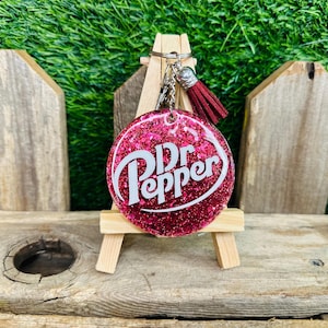 May include: A pink glitter resin keychain with a white Dr Pepper logo. The keychain is attached to a silver key ring with a brown tassel.