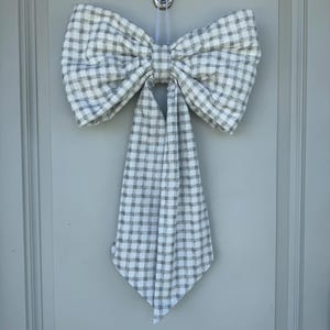 May include: A large, decorative bow made of gray and white checkered fabric, hanging on a light gray door. The bow is tied in the center and has long, flowing tails. The bow is attached to the door with a clear hook.