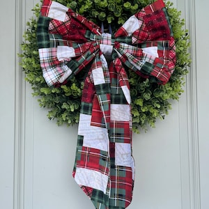May include: A round, green wreath with a large, decorative bow. The bow features a patchwork design in red, green, and white plaid patterns. The wreath is hanging on a white door, creating a festive holiday decoration.