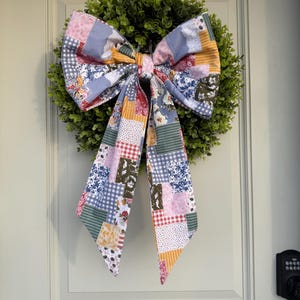 Può includere: Una ghirlanda decorativa con una base verde e fogliosa e un grande fiocco in tessuto patchwork. Il fiocco presenta una varietà di motivi e colori, tra cui stampe floreali, quadretti e righe. La ghirlanda è appesa a una porta bianca.