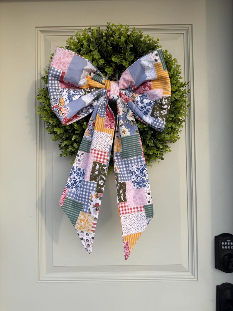 May include: A decorative wreath with a green, leafy base and a large, patchwork fabric bow. The bow features a variety of patterns and colors, including floral prints, gingham, and stripes. The wreath is hanging on a white door.