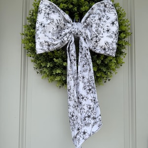 May include: A decorative wreath featuring a lush green base and a large bow. The bow is white with a black floral pattern. The wreath is designed for door or wall decor, adding a touch of elegance to any space.