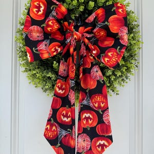 Puede incluir: Una corona de Halloween con una base circular verde y un gran lazo negro. El lazo está decorado con calabazas naranjas y telarañas. La corona está colgada en una puerta blanca.