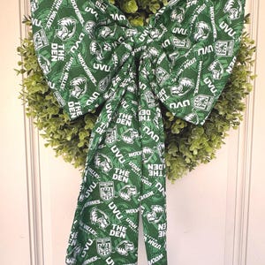 May include: A decorative wreath with a green bow. The bow is made of fabric printed with the text "UVU" and "Wolverines" in white on a green background. The wreath is surrounded by green foliage.