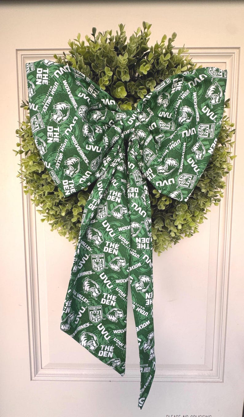 May include: A decorative wreath with a green bow. The bow is made of fabric printed with the text "UVU" and "Wolverines" in white on a green background. The wreath is surrounded by green foliage.