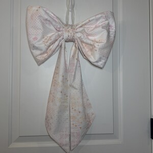 May include: A large, decorative fabric bow in shades of white, pink, and beige, hanging on a white door. The bow features a patterned design and is tied in the center, with two long, pointed tails hanging down. The bow is attached to the door with a clear adhesive hook.
