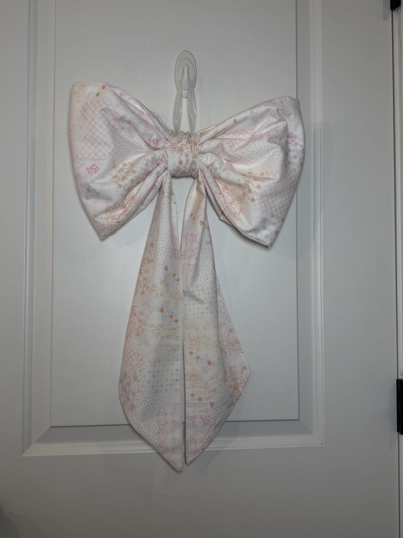 May include: A large, decorative fabric bow in shades of white, pink, and beige, hanging on a white door. The bow features a patterned design and is tied in the center, with two long, pointed tails hanging down. The bow is attached to the door with a clear adhesive hook.