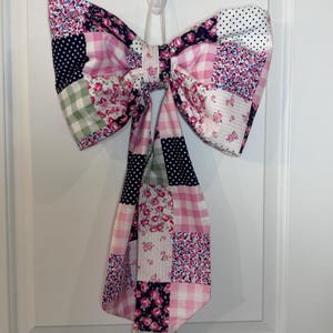 May include: A large, decorative bow made of patchwork fabric. The bow features a variety of patterns, including pink gingham, floral prints, and navy blue polka dots. It is hanging on a white door, adding a touch of charm to the decor.