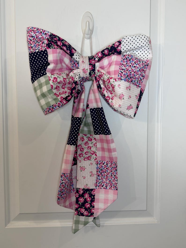 May include: A large, decorative bow made of patchwork fabric. The bow features a variety of patterns, including pink gingham, floral prints, and navy blue polka dots. It is hanging on a white door, adding a touch of charm to the decor.
