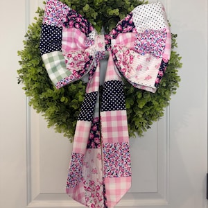May include: A decorative wreath with a large, patchwork bow. The bow features various fabric patterns, including polka dots, floral prints, and gingham in shades of pink, white, and navy blue. The wreath is made of green, leafy material.