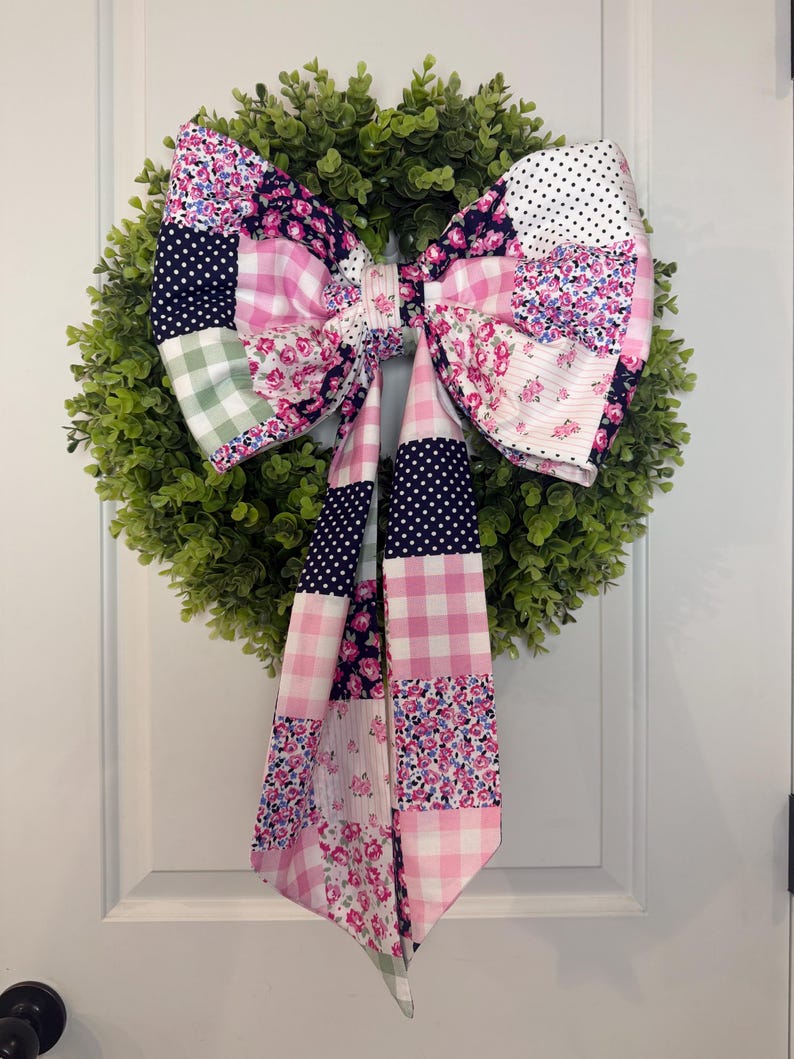May include: A decorative wreath with a large, patchwork bow. The bow features various fabric patterns, including polka dots, floral prints, and gingham in shades of pink, white, and navy blue. The wreath is made of green, leafy material.