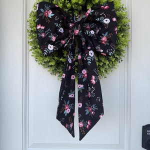 Puede incluir: Una corona verde con un gran lazo negro. El lazo está decorado con un estampado floral en tonos rosa, blanco y burdeos. La corona cuelga de una puerta blanca.