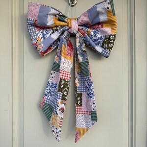May include: A large, decorative bow made of patchwork fabric, featuring a variety of floral, gingham, and solid patterns in shades of blue, pink, yellow, and green. The bow is hanging from a silver hook, with long, flowing tails.
