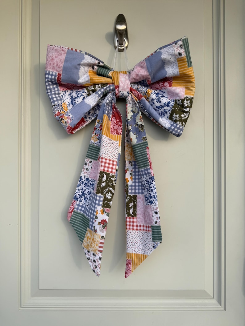 May include: A large, decorative bow made of patchwork fabric, featuring a variety of floral, gingham, and solid patterns in shades of blue, pink, yellow, and green. The bow is hanging from a silver hook, with long, flowing tails.